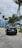 A black car parked on a sandy road with lush green palm trees and cloudy skies in the background.