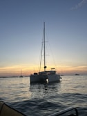 A catamaran is floating on calm waters during a sunset with a clear sky above. The horizon glows with shades of orange and pink, and a few other boats are visible in the distance.