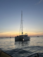 A catamaran is floating on calm waters during a sunset with a clear sky above. The horizon glows with shades of orange and pink, and a few other boats are visible in the distance.