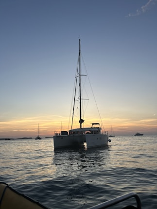A catamaran is floating on calm waters during a sunset with a clear sky above. The horizon glows with shades of orange and pink, and a few other boats are visible in the distance.