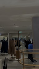 A modern, well-lit clothing store interior featuring several racks of hanging garments, including dresses and coats. The store has a minimalist design with neutral walls and a sleek tile floor. A central display holds a black high-heeled boot, adding a stylish touch. Mirrors are strategically placed to enhance the spacious feel.