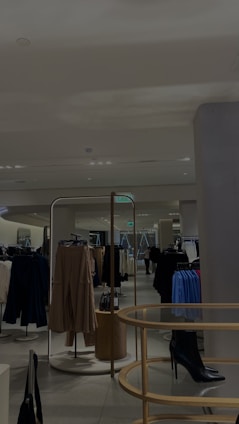 A modern, well-lit clothing store interior featuring several racks of hanging garments, including dresses and coats. The store has a minimalist design with neutral walls and a sleek tile floor. A central display holds a black high-heeled boot, adding a stylish touch. Mirrors are strategically placed to enhance the spacious feel.