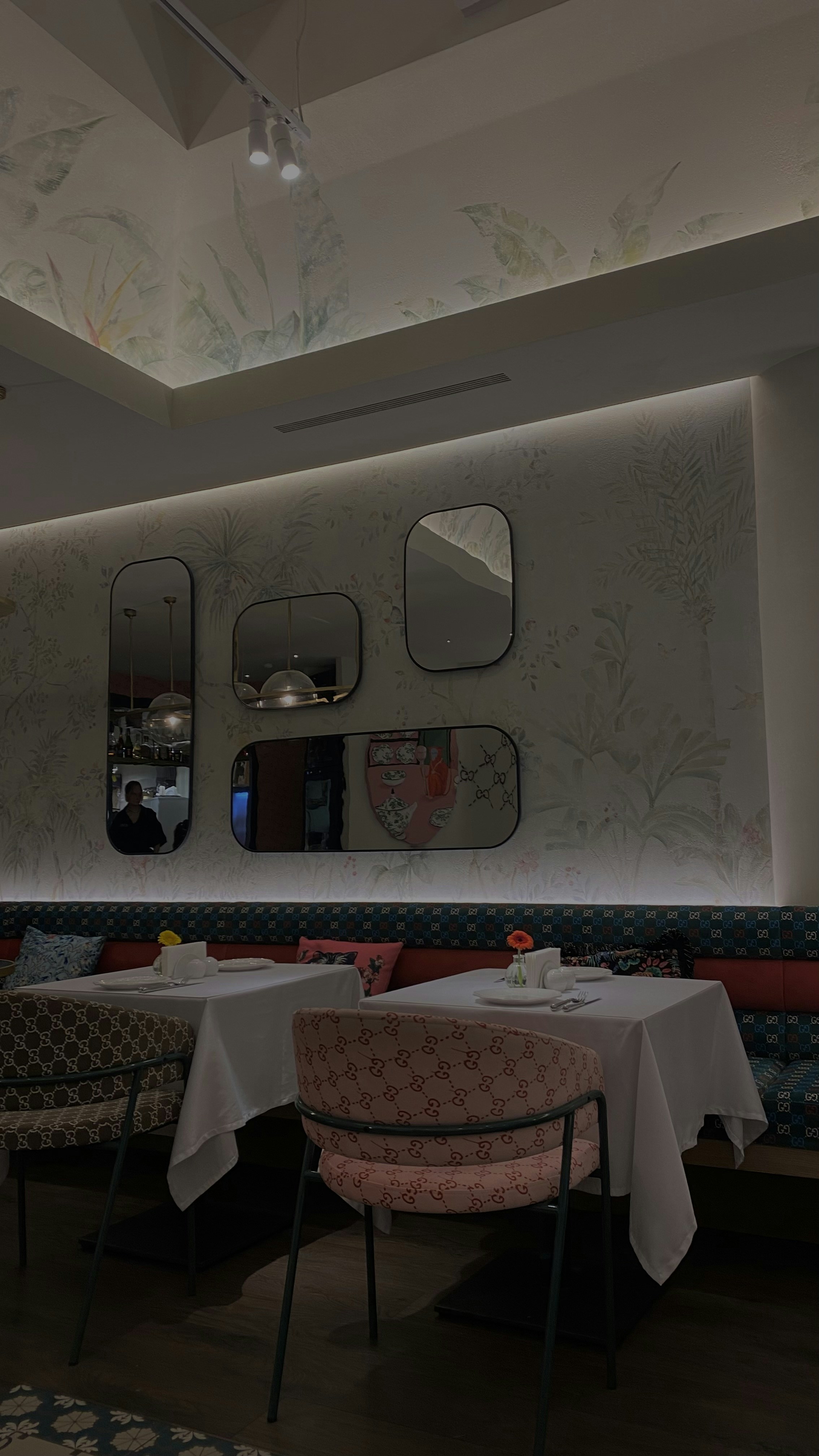 A modern, stylish restaurant interior featuring white tablecloth-covered tables with patterned chairs. Decorative mirrors are mounted on a wallpapered wall, and pendant lights hang from the ceiling. Each table is set with small vases holding colorful flowers, adding a touch of elegance.