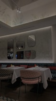 A modern, stylish restaurant interior featuring white tablecloth-covered tables with patterned chairs. Decorative mirrors are mounted on a wallpapered wall, and pendant lights hang from the ceiling. Each table is set with small vases holding colorful flowers, adding a touch of elegance.