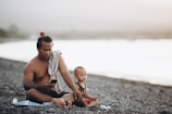 A man and a young child sit on a rocky beach. The man, shirtless and with a towel draped over one shoulder, is using a smartphone. The child, wearing a sleeveless shirt and red shorts, sits beside him holding a small toy vehicle. The background features a blurred view of water and a light, hazy sky.