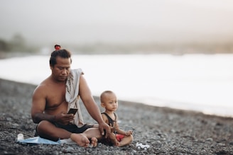 A man and a young child sit on a rocky beach. The man, shirtless and with a towel draped over one shoulder, is using a smartphone. The child, wearing a sleeveless shirt and red shorts, sits beside him holding a small toy vehicle. The background features a blurred view of water and a light, hazy sky.