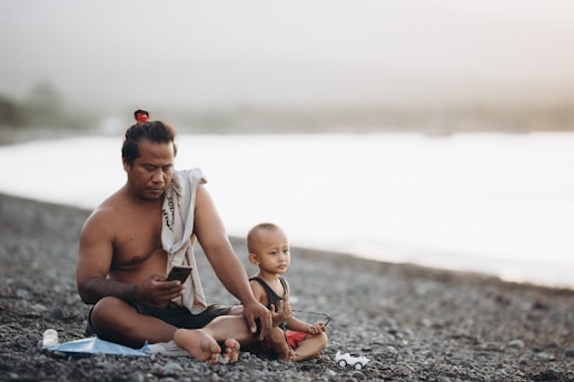 A man and a young child sit on a rocky beach. The man, shirtless and with a towel draped over one shoulder, is using a smartphone. The child, wearing a sleeveless shirt and red shorts, sits beside him holding a small toy vehicle. The background features a blurred view of water and a light, hazy sky.