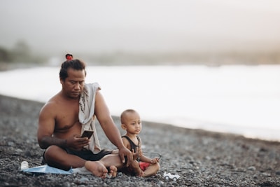 A man and a young child sit on a rocky beach. The man, shirtless and with a towel draped over one shoulder, is using a smartphone. The child, wearing a sleeveless shirt and red shorts, sits beside him holding a small toy vehicle. The background features a blurred view of water and a light, hazy sky.