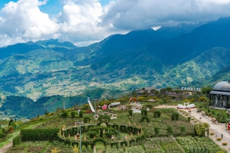 A picturesque landscape featuring lush green hills and mountains under a partly cloudy sky. The foreground showcases a manicured garden with neatly trimmed hedges, paths, and decorative elements, including sculptures and a dome-shaped structure. The mountainous backdrop adds depth and a sense of grandeur to the scene.