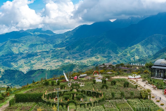A picturesque landscape featuring lush green hills and mountains under a partly cloudy sky. The foreground showcases a manicured garden with neatly trimmed hedges, paths, and decorative elements, including sculptures and a dome-shaped structure. The mountainous backdrop adds depth and a sense of grandeur to the scene.