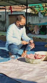 A person wearing a mask is preparing meat, possibly cutting it into smaller pieces. The setting appears to be outdoors under a canopy, with additional people seated on the ground in the background. There is a large bowl containing meat in front of the person, and other materials laid out on the ground.