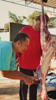 Two men are involved in the process of cutting meat from an animal carcass. One man, wearing a blue shirt, is focused on using a knife to separate the meat. The other man is in a red shirt, holding up the carcass for support. They are under a covered area, with trees visible in the background.