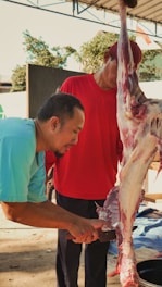 Two men are involved in the process of cutting meat from an animal carcass. One man, wearing a blue shirt, is focused on using a knife to separate the meat. The other man is in a red shirt, holding up the carcass for support. They are under a covered area, with trees visible in the background.