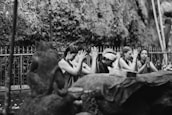 A group of people engaged in a spiritual or meditative practice, sitting in a row with hands raised in prayer or meditation gestures. The setting appears to be outdoors with a natural backdrop and metal fencing. The atmosphere is serene and focused.