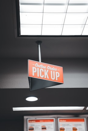 A red sign hangs from the ceiling, indicating the area for online order pickup. The sign is triangular with white text, and nearby, illuminated menus display food items.
