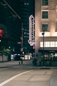 A nighttime view of a city street featuring a hotel sign on the side of a building. The illuminated Lancaster Hotel sign is prominent, with the street below appearing quiet and empty. Nearby buildings are mostly dark, with some windows lit up.
