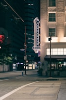 A nighttime view of a city street featuring a hotel sign on the side of a building. The illuminated Lancaster Hotel sign is prominent, with the street below appearing quiet and empty. Nearby buildings are mostly dark, with some windows lit up.