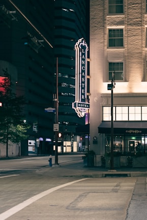 A nighttime view of a city street featuring a hotel sign on the side of a building. The illuminated Lancaster Hotel sign is prominent, with the street below appearing quiet and empty. Nearby buildings are mostly dark, with some windows lit up.