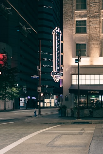 A nighttime view of a city street featuring a hotel sign on the side of a building. The illuminated Lancaster Hotel sign is prominent, with the street below appearing quiet and empty. Nearby buildings are mostly dark, with some windows lit up.