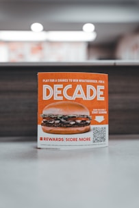 A promotional sign displaying a burger with layers of lettuce, tomato, and beef patty. The sign features the word 'Decade' in bold letters, promoting a contest with a QR code for participation. The background is softly blurred, highlighting the sign as the main focus.