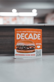 A promotional sign displaying a burger with layers of lettuce, tomato, and beef patty. The sign features the word 'Decade' in bold letters, promoting a contest with a QR code for participation. The background is softly blurred, highlighting the sign as the main focus.