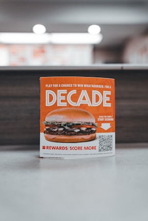 A promotional sign displaying a burger with layers of lettuce, tomato, and beef patty. The sign features the word 'Decade' in bold letters, promoting a contest with a QR code for participation. The background is softly blurred, highlighting the sign as the main focus.