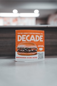 A promotional sign displaying a burger with layers of lettuce, tomato, and beef patty. The sign features the word 'Decade' in bold letters, promoting a contest with a QR code for participation. The background is softly blurred, highlighting the sign as the main focus.