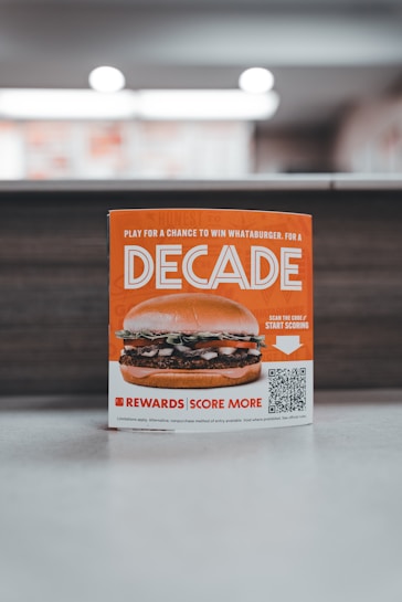 A promotional sign displaying a burger with layers of lettuce, tomato, and beef patty. The sign features the word 'Decade' in bold letters, promoting a contest with a QR code for participation. The background is softly blurred, highlighting the sign as the main focus.
