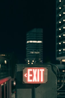 An illuminated red EXIT sign mounted on a concrete wall, surrounded by a dark urban environment. In the background, there are blurry lights from a high-rise building and other city elements, emphasizing a nighttime setting.