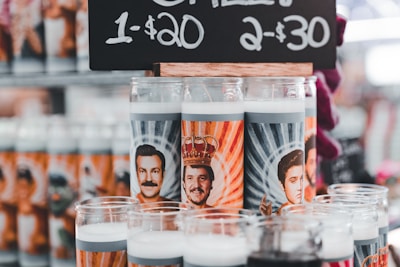 A variety of novelty prayer candles featuring illustrations of well-known figures with stylized backgrounds. They are displayed under a price sign offering them for sale with a special deal.