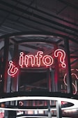 A neon sign displaying the word 'info' with a question mark, set against a backdrop of industrial-style architecture. The sign is enclosed in a wireframe structure and illuminated with red and white neon lights.