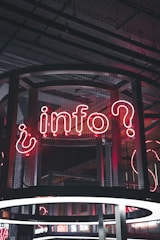 A neon sign displaying the word 'info' with a question mark, set against a backdrop of industrial-style architecture. The sign is enclosed in a wireframe structure and illuminated with red and white neon lights.