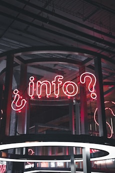 A neon sign displaying the word 'info' with a question mark, set against a backdrop of industrial-style architecture. The sign is enclosed in a wireframe structure and illuminated with red and white neon lights.