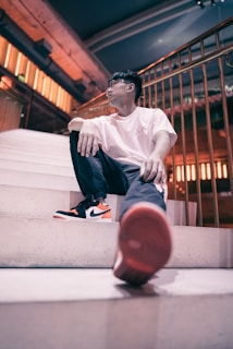A person sits casually on a set of wide stairs, leaning back comfortably. The individual is dressed in a white t-shirt, dark pants, and black and orange sneakers. The setting suggests a modern, urban environment with architectural elements like a metal railing and warm lighting, casting a relaxed and stylish vibe.