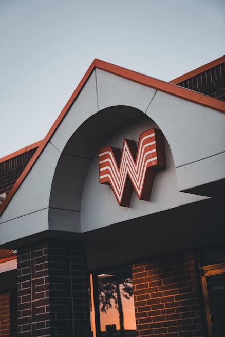 A building with a distinctive architectural design features a large orange and white stylized letter W logo under an arch. The structure is made of brick and stucco, and the lighting suggests it is either sunrise or sunset.