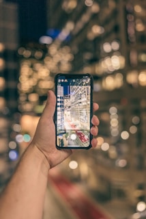 A hand holding a smartphone, capturing a photograph of a brightly lit city street at night. The background of the image is out of focus, creating a bokeh effect with the city lights.