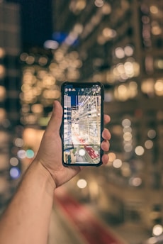 A hand holding a smartphone, capturing a photograph of a brightly lit city street at night. The background of the image is out of focus, creating a bokeh effect with the city lights.