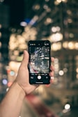 A hand is holding a smartphone with the camera app open, showing a live view of a cityscape featuring tall buildings with lights. The background is blurred, creating a bokeh effect of city lights.