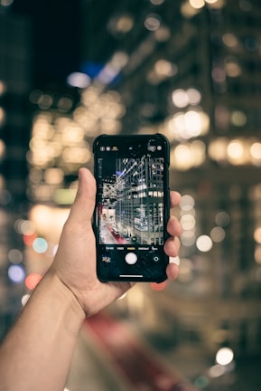 A hand is holding a smartphone with the camera app open, showing a live view of a cityscape featuring tall buildings with lights. The background is blurred, creating a bokeh effect of city lights.