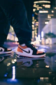 Sneakers with vibrant orange detailing stepping on a wet street reflecting neon signs.