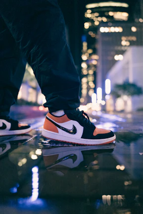 Close-up of chunky sneakers stepping on wet pavement reflecting city lights in charcoal and purple hues.