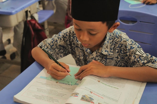 A young boy is focused on writing notes while sitting at a classroom desk. He is wearing a patterned shirt and a traditional cap. The boy rests his left hand on an open textbook, suggesting an educational setting.