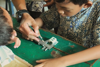 A young boy intently watches as an adult, wearing a watch, demonstrates how to use a green mechanical pencil on a metallic tool placed on an old green table. Another child is observing closely from the side.