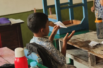 A young boy is sitting at a school desk, holding a paper airplane in his hand. The classroom appears simple, with a wooden chair inverted on the desk next to him and a water bottle visible nearby. The boy is wearing a patterned shirt and has a backpack on the back of his chair.