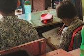 Two children in patterned shirts are seated at wooden desks. One is drawing in a notebook with a pencil, focusing intently on the task. A blue water bottle and a red pencil case are on the desk.