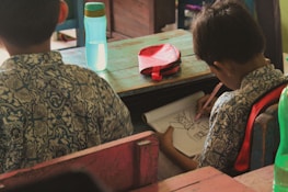 Two children in patterned shirts are seated at wooden desks. One is drawing in a notebook with a pencil, focusing intently on the task. A blue water bottle and a red pencil case are on the desk.