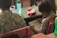 Two children in patterned shirts are seated at wooden desks. One is drawing in a notebook with a pencil, focusing intently on the task. A blue water bottle and a red pencil case are on the desk.