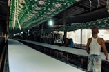 A long, narrow factory-like setting with two men working. They are handling large rolls of fabric on an extended table. The area is covered with green cloth decorated with patterns, hanging overhead. Strip lighting illuminates the space, creating shadows and highlights.