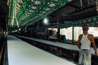 A long, narrow factory-like setting with two men working. They are handling large rolls of fabric on an extended table. The area is covered with green cloth decorated with patterns, hanging overhead. Strip lighting illuminates the space, creating shadows and highlights.