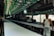 A long, narrow factory-like setting with two men working. They are handling large rolls of fabric on an extended table. The area is covered with green cloth decorated with patterns, hanging overhead. Strip lighting illuminates the space, creating shadows and highlights.